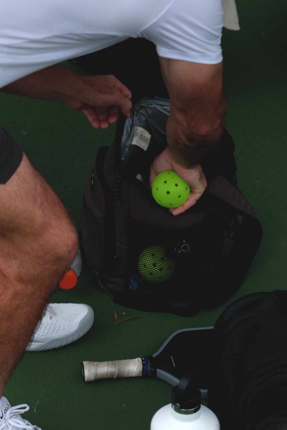 Person placing a pickleball into a backpack on a court.