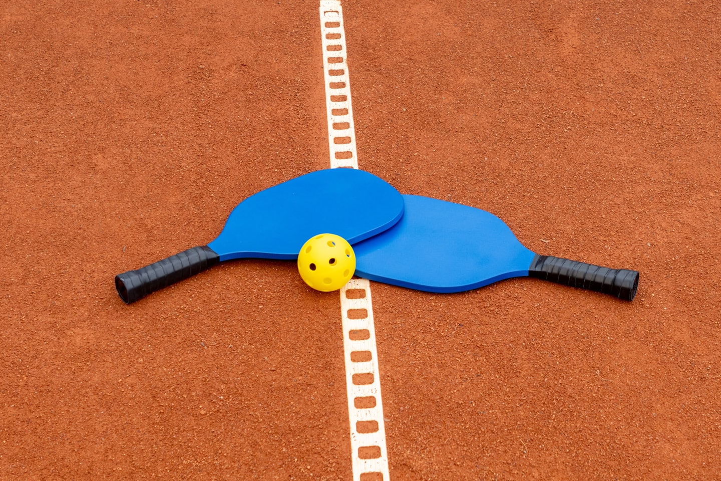 Two blue pickleball paddles and a yellow ball on a court.