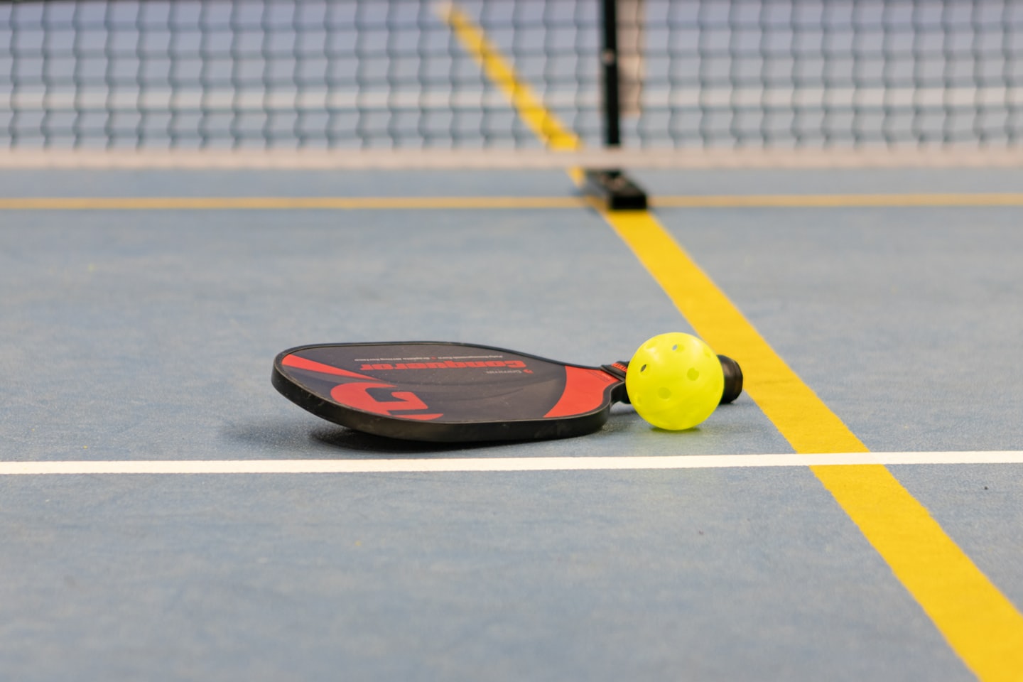 A black and red pickleball paddle and yellow ball on a blue court.