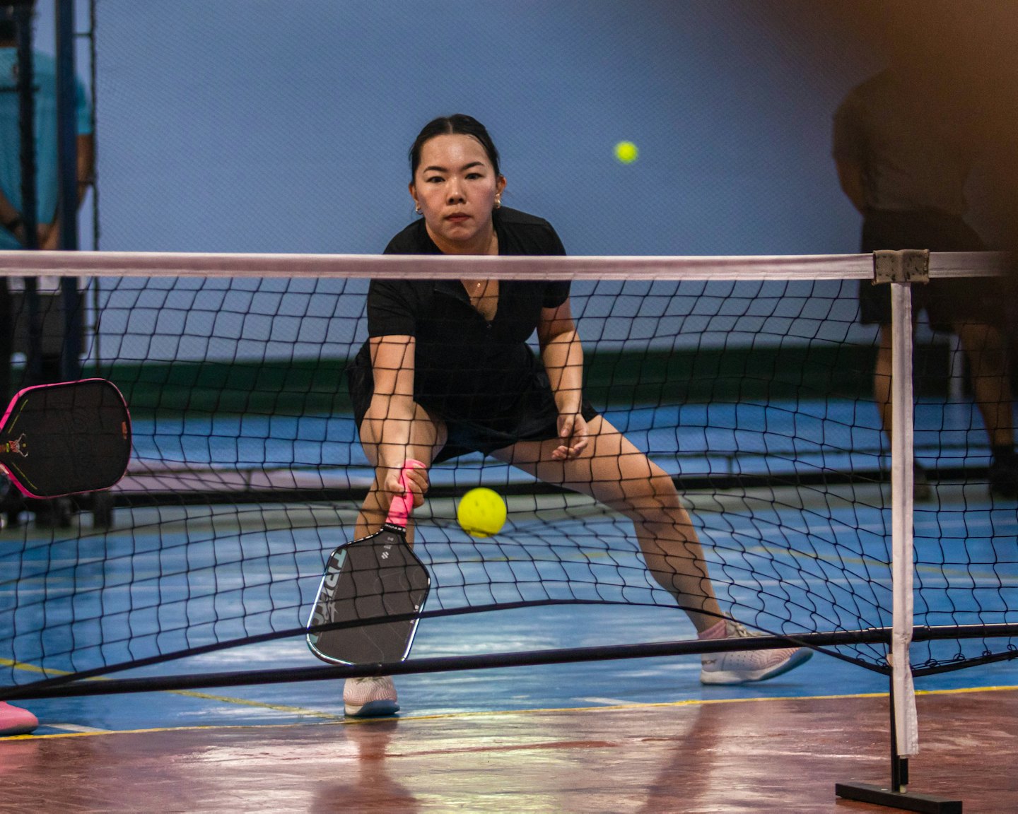 Woman crouches low while playing pickleball on an indoor court.