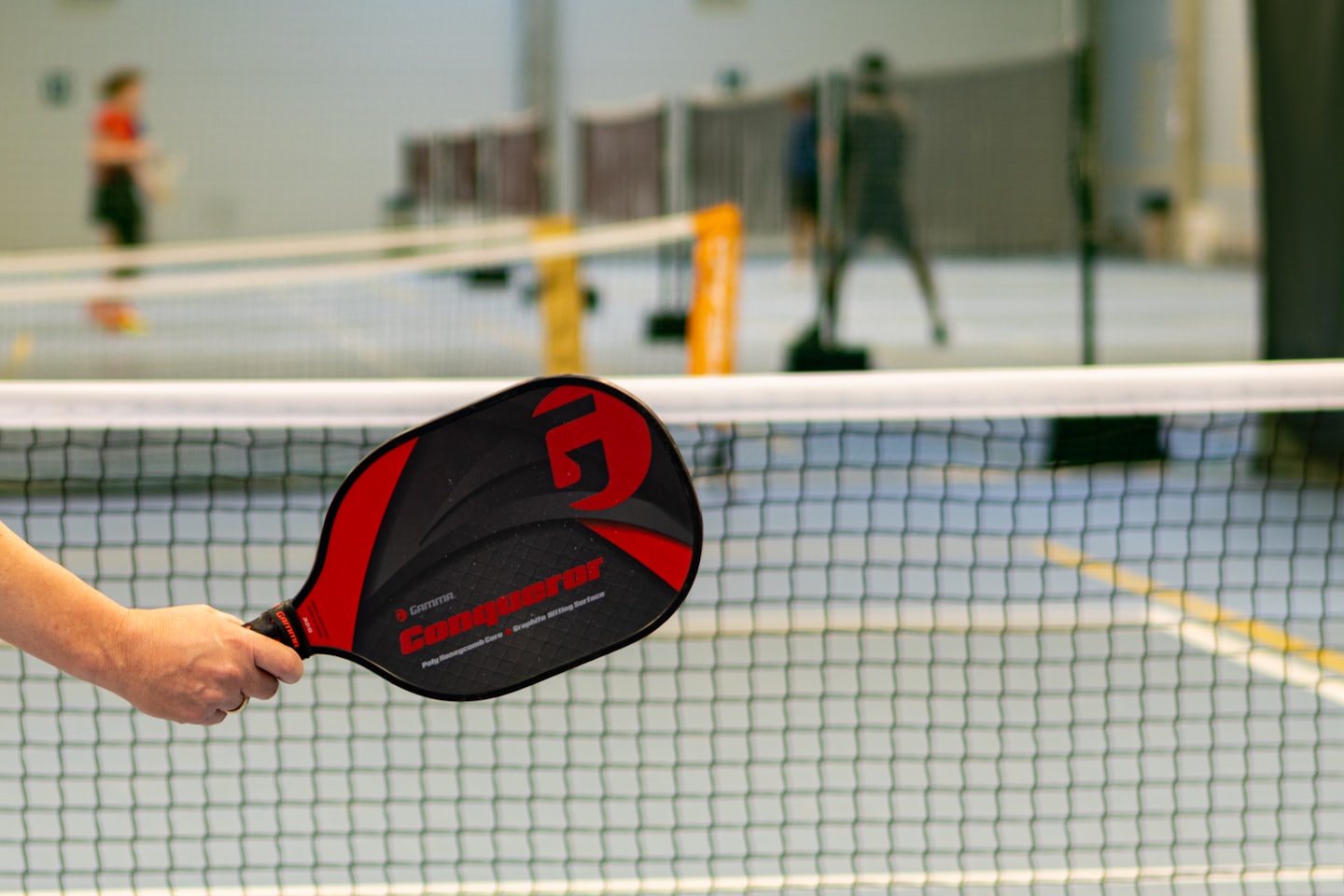 A hand grips a pickleball paddle over the indoor court net.
